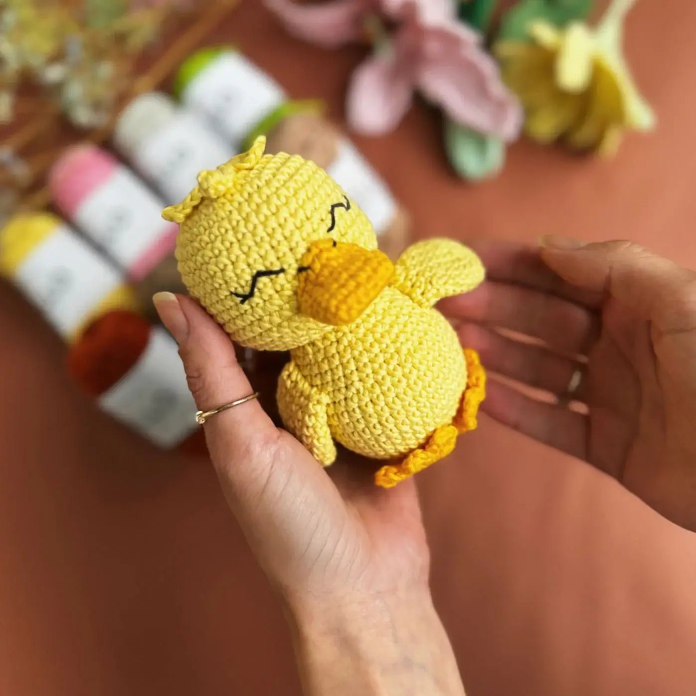 Hand holding a small yellow crocheted duck toy with a blurred background of balls of cotton yarn and flowers.
