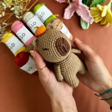Hand holding a small brown crocheted capybara toy with yarn and flowers in the background.