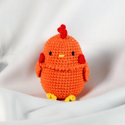 Crocheted orange chicken toy with red accent feathers on a white fabric background