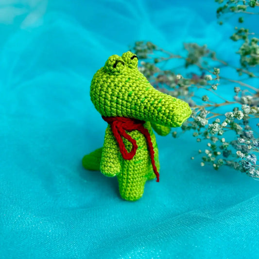 Crocheted green crocodile standing up with a red scarf on a turquoise background