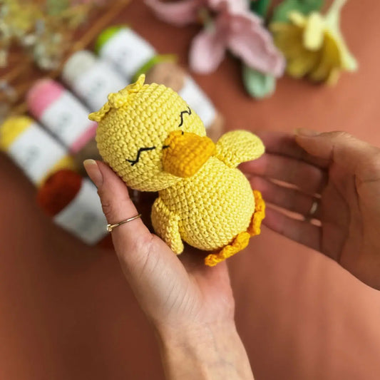 Hand holding a small yellow crocheted duck toy with a blurred background of balls of cotton yarn and flowers.