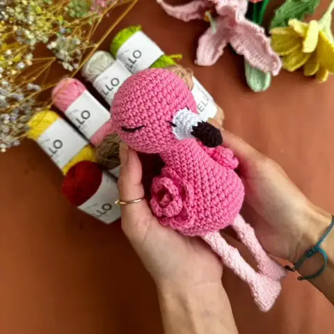 Hand holding a pink crochet toy with flowers and yarn in the background
