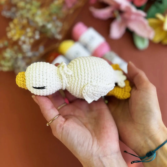 Hand holding a small crocheted toy  a white goose with a blurred floral background