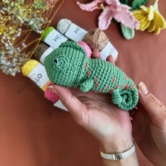 Hand holding a green crocheted chameleon toy with yarn and flowers in the background