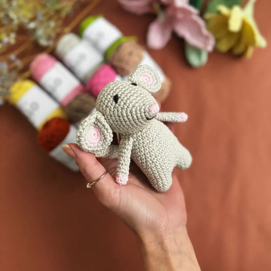 Hand holding a small crocheted mouse toy with flowers and yarn in the background