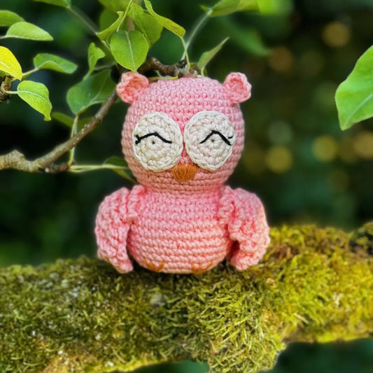 Pink crochet owl sitting on a branch with green leaves in the background