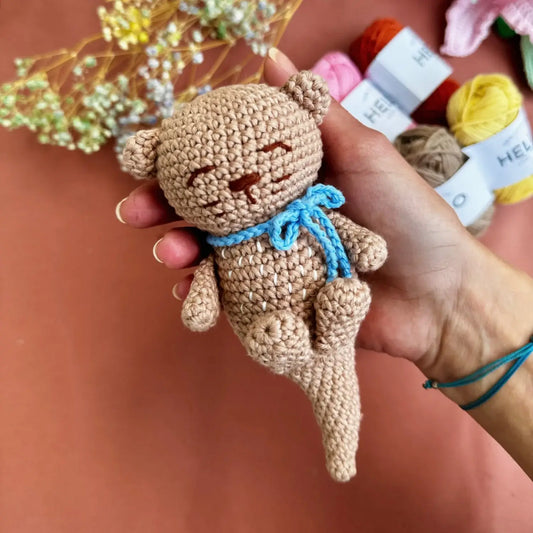 Hand holding a small crocheted otter toy with yarn and flowers in the background