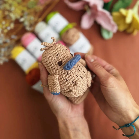Hand holding a small crocheted Platypus animal toy with a blurred background of craft supplies.