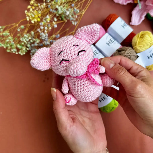 Hand holding a pink crochet pig toy who has a pink bow, with yarn and flowers in the background