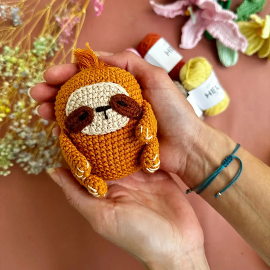 Hand holding a small crochet sloth toy with yarn and flowers in the background